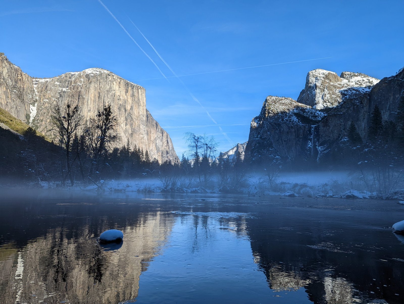 Granite cliffs of Yosemite National Park tower over the misty Merced river, reflecting in the water.